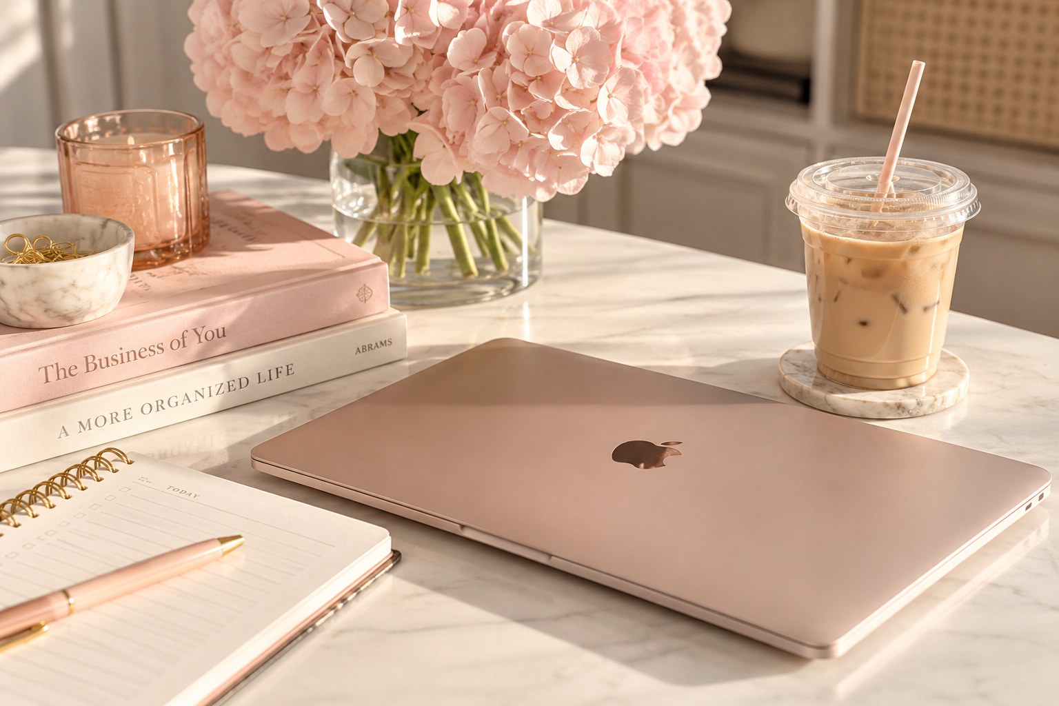 Closed rose-gold laptop and iced latte on a softly lit blush workspace